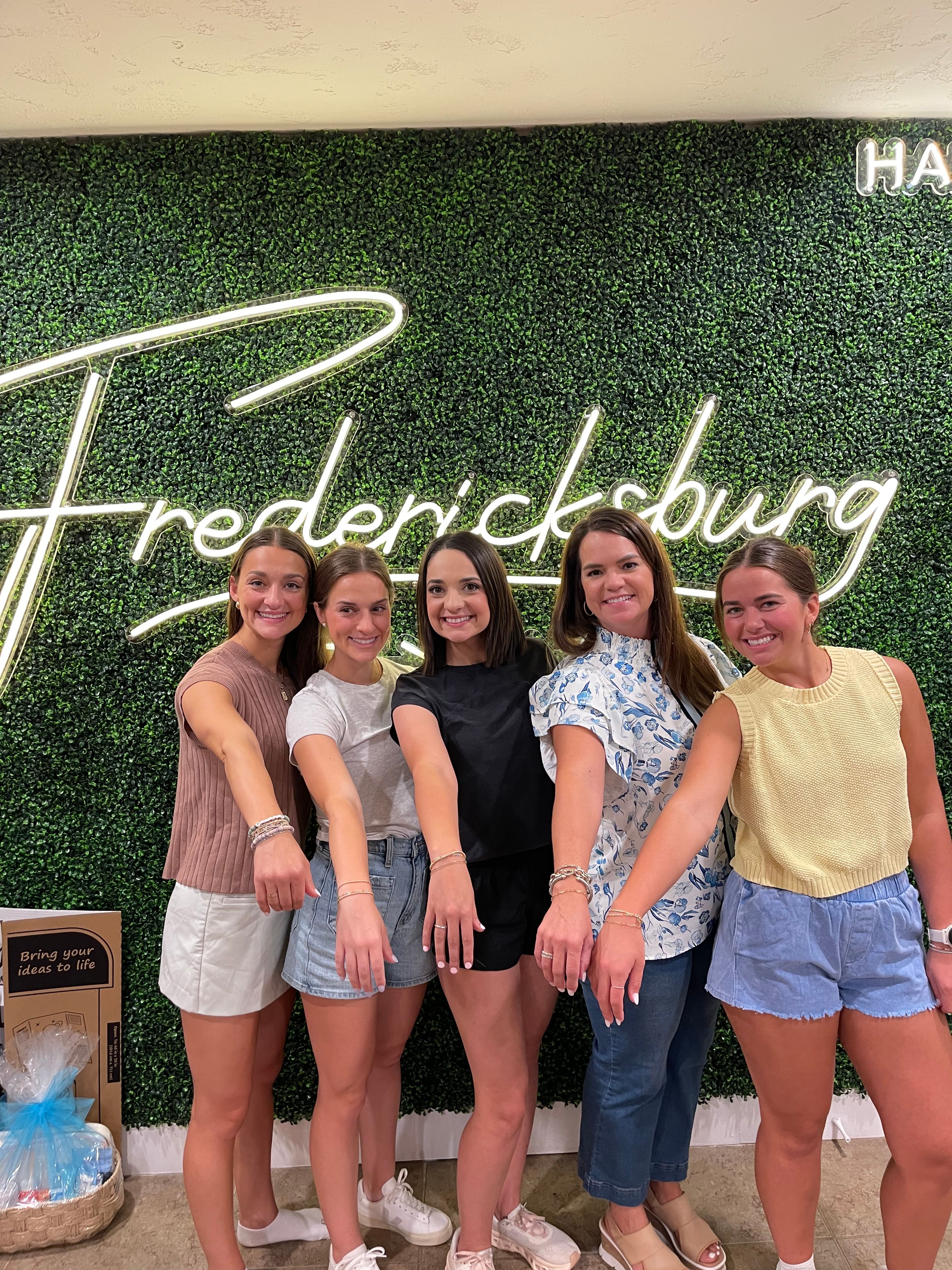 Five smiling women posing in front of a green hedge wall with neon 'Fredericksburg' script, extending their arms to show matching bracelets — fun group photo.