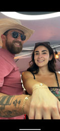 Smiling couple selfie under a pink canopy at an outdoor market — man in a straw cowboy hat, sunglasses and tattooed arm; woman in a floral top showing bracelets with string light overhead