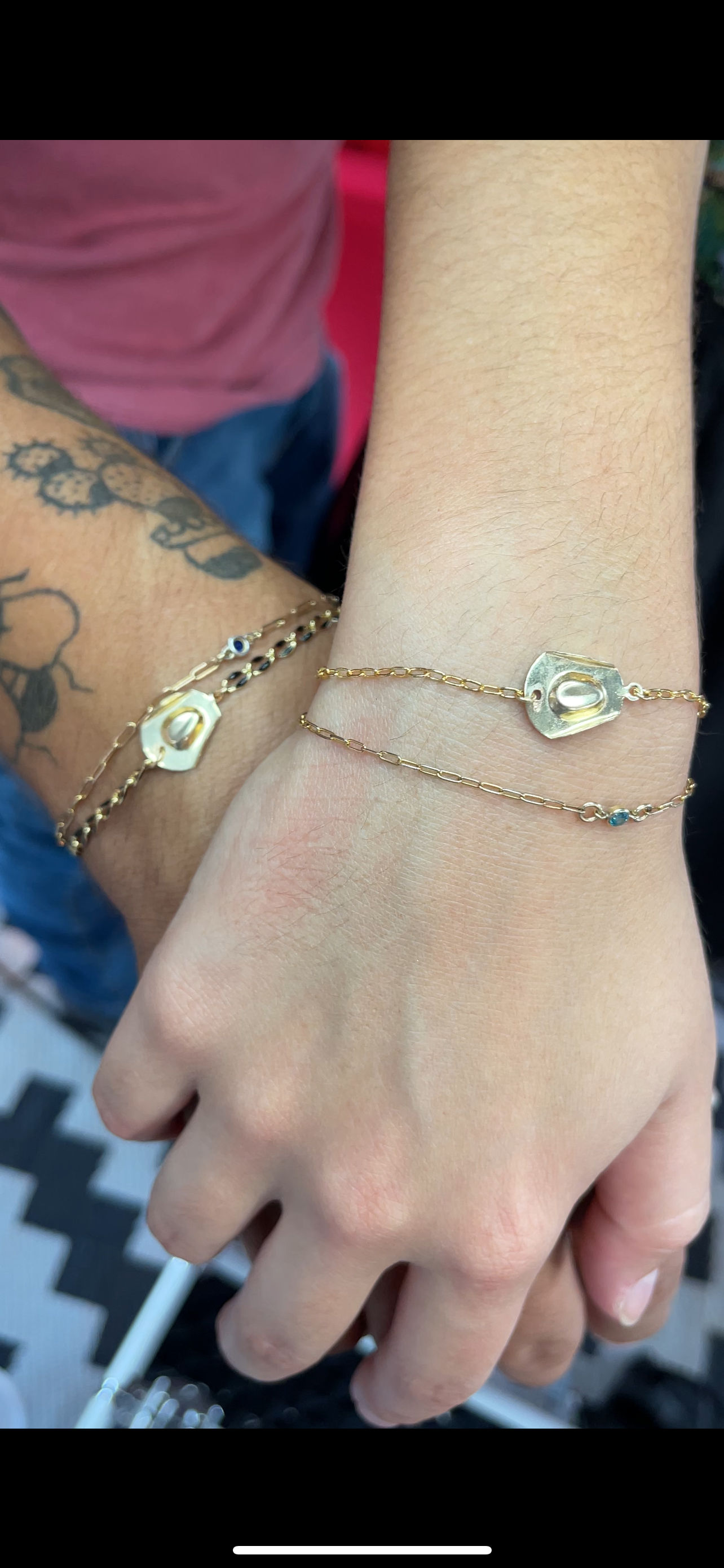 Close-up of two hands wearing matching delicate gold chain bracelets with organic-shaped gold charms and tiny blue stones—stacked minimalist jewelry shot.