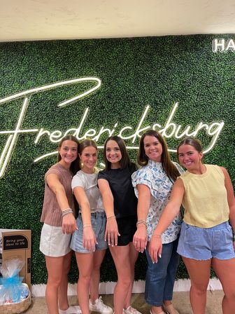 Five smiling women in casual summer outfits showing matching bracelets while posing in front of a green hedge wall with a neon "Fredericksburg" sign
