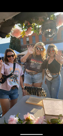 Three smiling women in sunglasses waving at an outdoor street fair jewelry booth under pink flower garlands and a hanging disco ball on a sunny summer day.