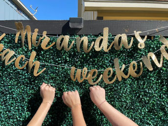 Three raised fists wearing matching string bracelets at an outdoor backyard celebration against a faux boxwood wall and a sparkling gold banner that reads "weekend".