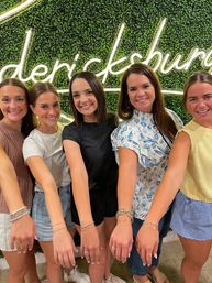 Five smiling women in casual summer outfits posing indoors in front of a green boxwood wall with a neon cursive sign, showing off matching delicate bracelets on their outstretched wrists.