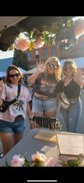 Three smiling women waving at an outdoor downtown street fair vendor booth, posing behind a table of dainty bracelets and pink flower garlands under a hanging disco ball on a sunny summer afternoon.