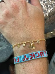 Close-up wrist jewelry: delicate gold charm chain with tiny cowboy hat, bird and letter charms paired with a turquoise and red beaded bracelet, photographed against a sparkly sequined background.