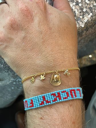 Close-up wrist jewelry: delicate gold charm chain with tiny cowboy hat, bird and letter charms paired with a turquoise and red beaded bracelet, photographed against a sparkly sequined background.