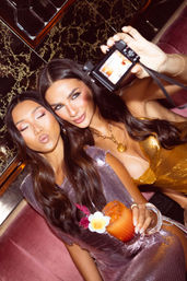 Two friends taking a glamorous nightlife selfie in a velvet lounge, wearing sequined dresses; one holds a compact camera while the other holds a tropical cocktail garnished with a flower.