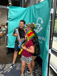 Group posing in an indoor photo booth with a teal backdrop, smiling and holding inflatable instruments — keyboard, guitar and toy microphone — child wearing a rainbow wig and checkered shorts at a casual party event.