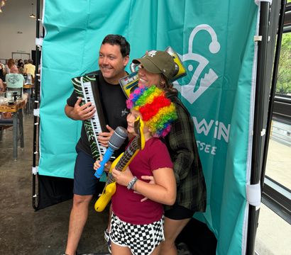 Group posing in an indoor photo booth with a teal backdrop, smiling and holding inflatable instruments — keyboard, guitar and toy microphone — child wearing a rainbow wig and checkered shorts at a casual party event.