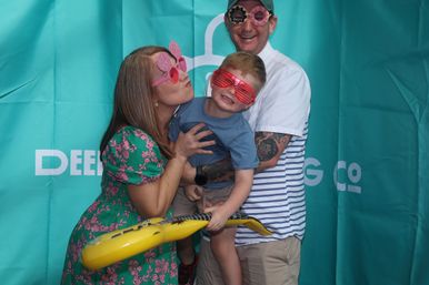 Family of three in a photo-booth style shot against a teal backdrop — parents wearing playful novelty sunglasses hold a smiling child in red shutter shades clutching an inflatable yellow guitar.