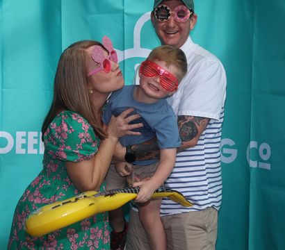 Family of three in a photo-booth style shot against a teal backdrop — parents wearing playful novelty sunglasses hold a smiling child in red shutter shades clutching an inflatable yellow guitar.