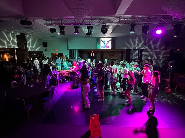 Indoor dance party in an event hall — crowd dancing on a glossy floor under purple and teal stage lights with overhead lighting rig and a wet-floor caution sign.
