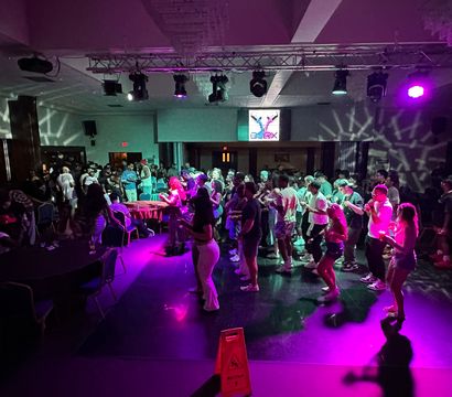 Indoor dance party in an event hall — crowd dancing on a glossy floor under purple and teal stage lights with overhead lighting rig and a wet-floor caution sign.