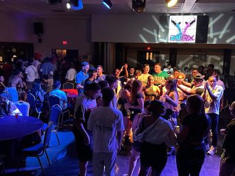 Crowded indoor nightclub scene with a lively dance floor, colorful stage lights and spotlights, a large video screen showing a vibrant pill-shaped graphic, and people socializing around tables.