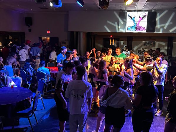Crowded indoor nightclub scene with a lively dance floor, colorful stage lights and spotlights, a large video screen showing a vibrant pill-shaped graphic, and people socializing around tables.