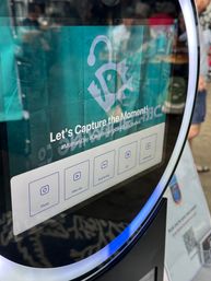 Interactive selfie kiosk touchscreen with teal backdrop and “Let’s Capture the Moment!” prompt, Photo/Slow‑mo/Boomerang/GIF icons, glowing LED ring and faint reflections of event attendees.