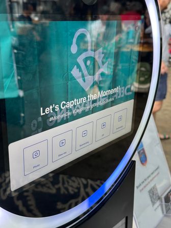 Interactive selfie kiosk touchscreen with teal backdrop and “Let’s Capture the Moment!” prompt, Photo/Slow‑mo/Boomerang/GIF icons, glowing LED ring and faint reflections of event attendees.