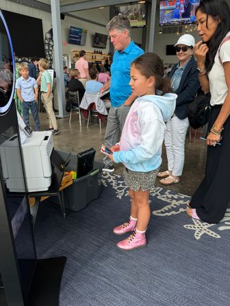 Girl in pastel hoodie and pink boots using a photo booth at a busy family-friendly indoor venue with a bar and TV screens