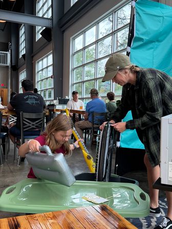 Adult helping a child with inflatable pool toys in a bright, family-friendly industrial cafe with floor-to-ceiling windows and wooden tables.