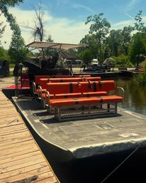 Bright orange-seat airboat with twin rear fan cages docked at a wooden pier in a sunny bayou surrounded by cypress trees, ready for a swamp tour.