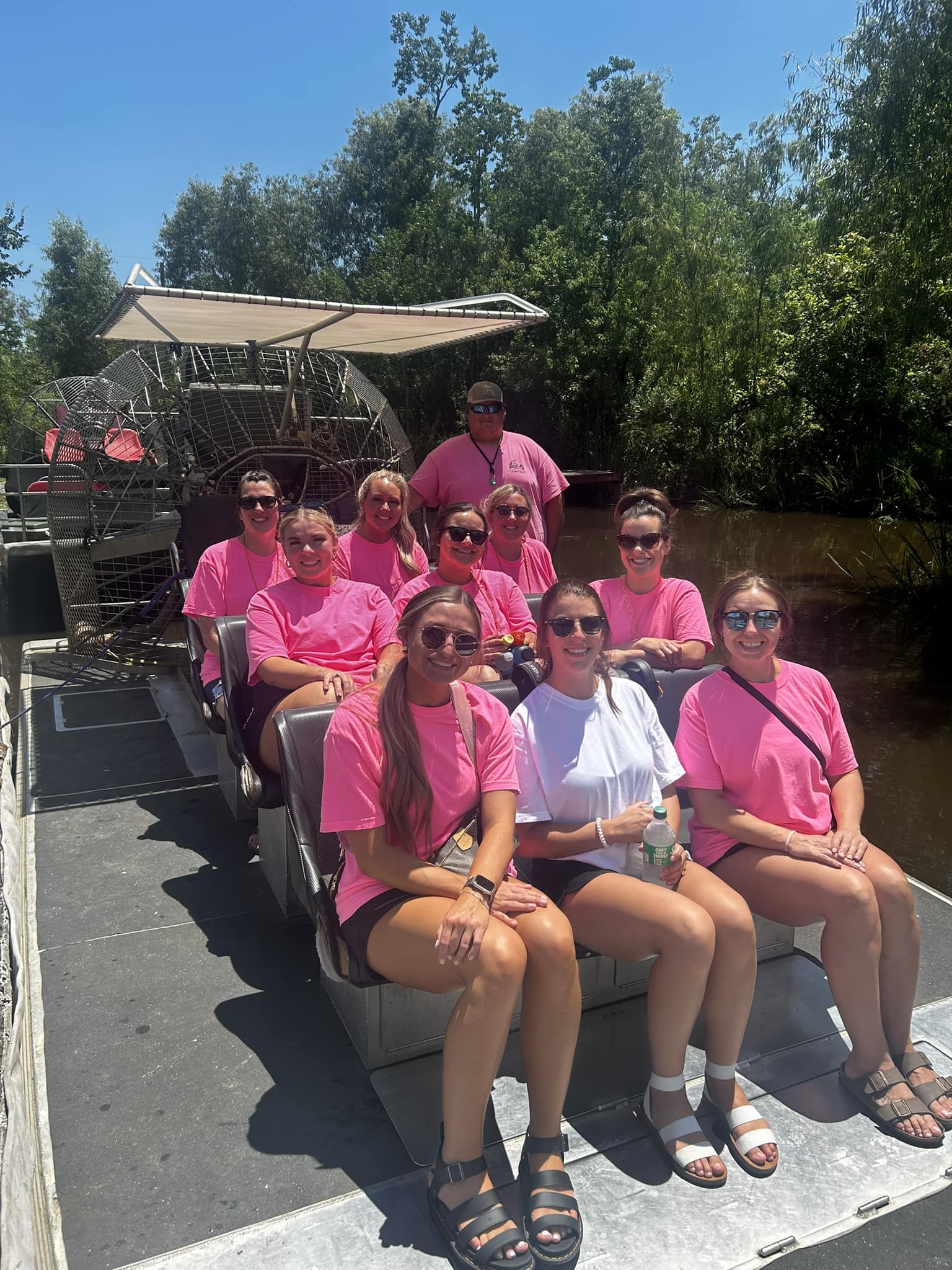Group of friends in matching pink shirts smiling on an airboat tour through sunlit swamp wetlands under a clear blue sky