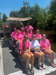 Group of friends in matching pink shirts smiling on an airboat tour through sunlit swamp wetlands under a clear blue sky