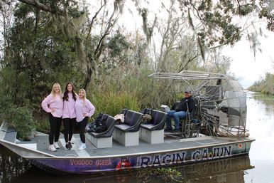 Three women in matching pink shirts pose on the bow of an airboat during a Louisiana bayou airboat tour, operator seated by the rear fan, surrounded by cypress trees draped with Spanish moss and calm swamp water.