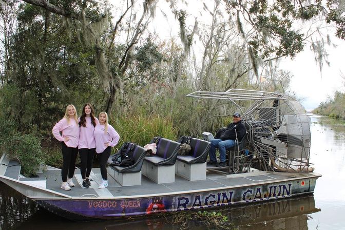 Three women in matching pink shirts pose on the bow of an airboat during a Louisiana bayou airboat tour, operator seated by the rear fan, surrounded by cypress trees draped with Spanish moss and calm swamp water.
