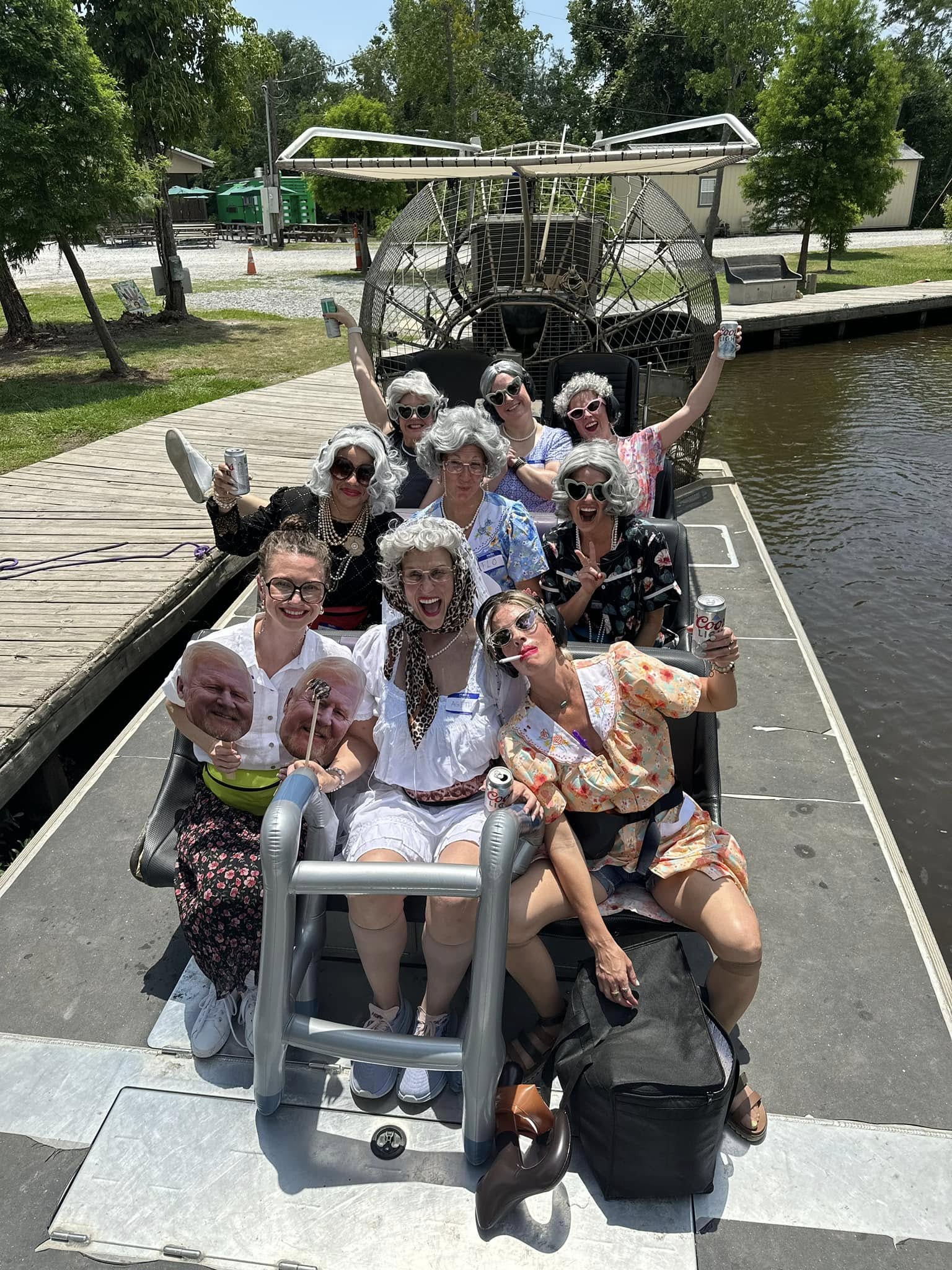 Group of people in silver wigs and retro outfits laughing and holding drinks on an airboat at a riverside dock on a sunny day