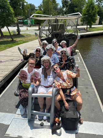 Group of people in silver wigs and retro outfits laughing and holding drinks on an airboat at a riverside dock on a sunny day