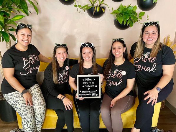 Five smiling women in matching 'Bridal Squad' tees and heart-shaped sunglasses sit on a yellow couch holding a letterboard reading bride-to-be, with hanging plants behind — playful bachelorette party photo