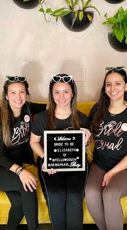 Five smiling women in matching 'Bridal Squad' tees and heart-shaped sunglasses sit on a yellow couch holding a letterboard reading bride-to-be, with hanging plants behind — playful bachelorette party photo