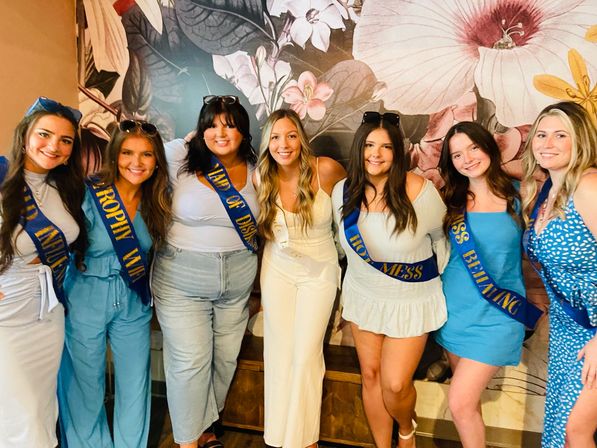 Seven smiling women posing arm-in-arm indoors in front of a large floral mural; bride-to-be in white surrounded by friends in blue outfits wearing celebratory sashes for a bachelorette party