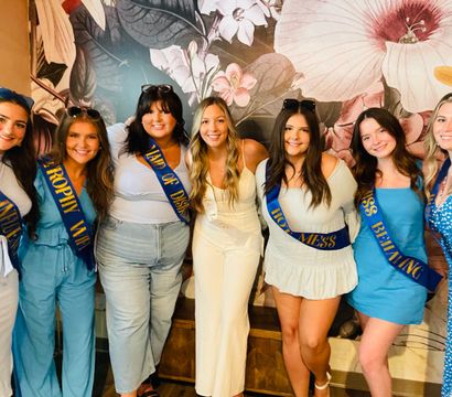 Seven smiling women posing arm-in-arm indoors in front of a large floral mural; bride-to-be in white surrounded by friends in blue outfits wearing celebratory sashes for a bachelorette party