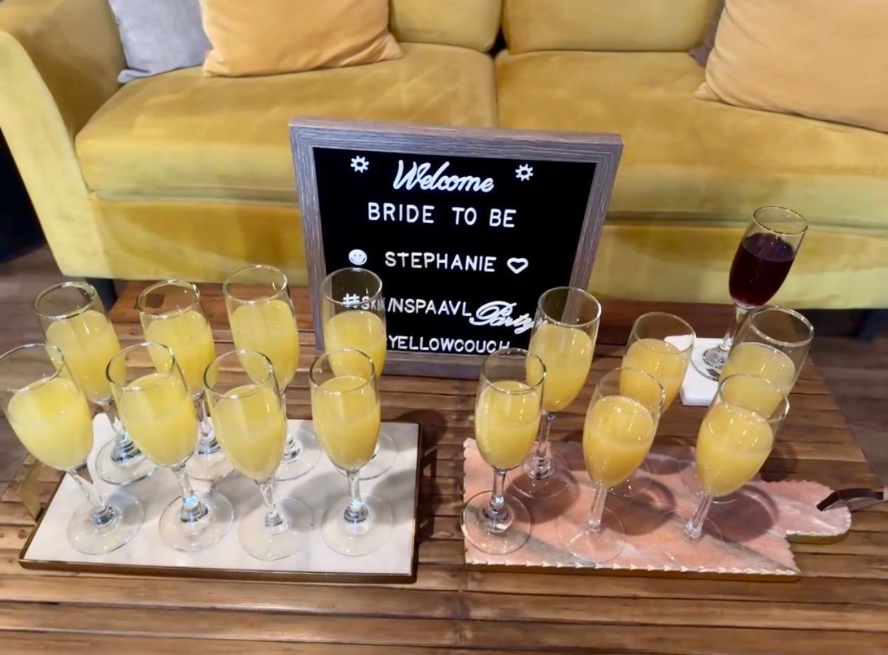 Mimosa-filled champagne flutes arranged on trays around a small welcome sign for a bride-to-be, set on a wooden coffee table in front of a yellow couch — cozy bachelorette brunch scene.
