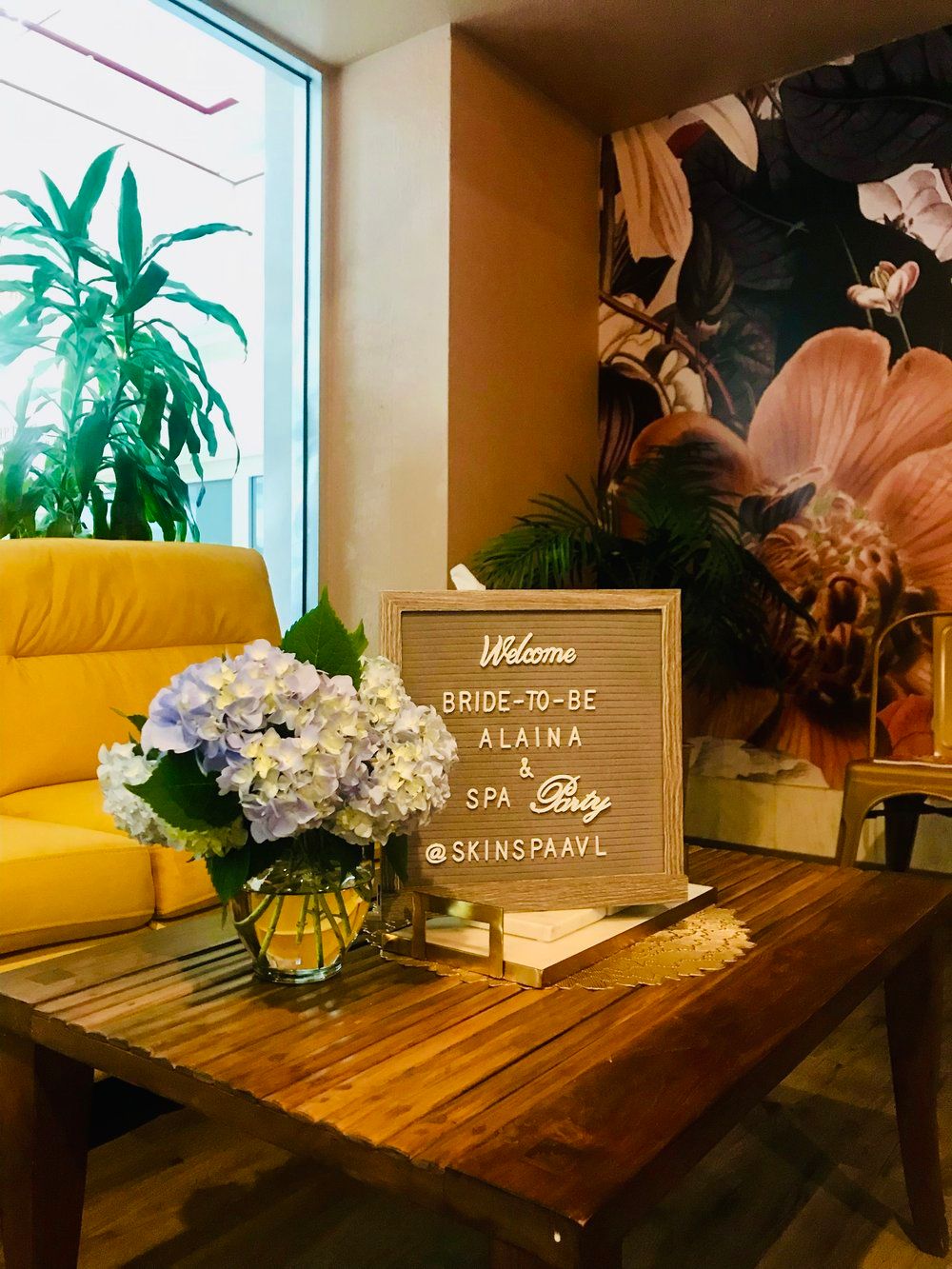 Boutique spa lounge with wooden coffee table, pastel hydrangea bouquet and a framed letterboard reading “Welcome Bride‑to‑Be & Spa Party” beside a yellow armchair and tropical mural