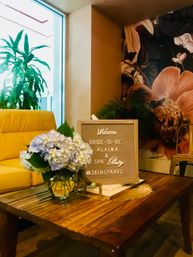 Boutique spa lounge with wooden coffee table, pastel hydrangea bouquet and a framed letterboard reading “Welcome Bride‑to‑Be & Spa Party” beside a yellow armchair and tropical mural
