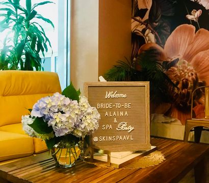 Boutique spa lounge with wooden coffee table, pastel hydrangea bouquet and a framed letterboard reading “Welcome Bride‑to‑Be & Spa Party” beside a yellow armchair and tropical mural