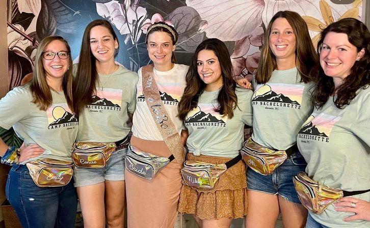 Six women in matching 'Camp Bachelorette' tees and holographic fanny packs pose indoors for a bachelorette photo; bride-to-be wears a sash and 'Bride' pouch — camp-themed party in Asheville, NC.