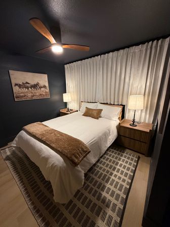 Cozy modern bedroom interior with a white-dressed bed and brown throw, wooden nightstands and lamps, floor-to-ceiling beige curtains, patterned rug, wooden-blade ceiling fan, and horse wall art on a dark accent wall.