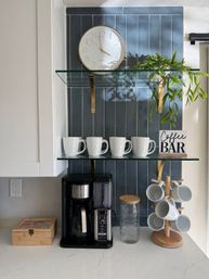 Cozy kitchen coffee station with navy vertical tile backsplash, round wall clock, glass shelves holding four white mugs, trailing green plant and a "Coffee Bar" sign above a black drip coffee maker and wooden mug tree on a white quartz countertop.