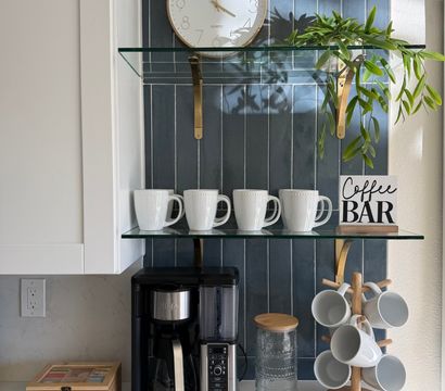 Cozy kitchen coffee station with navy vertical tile backsplash, round wall clock, glass shelves holding four white mugs, trailing green plant and a "Coffee Bar" sign above a black drip coffee maker and wooden mug tree on a white quartz countertop.