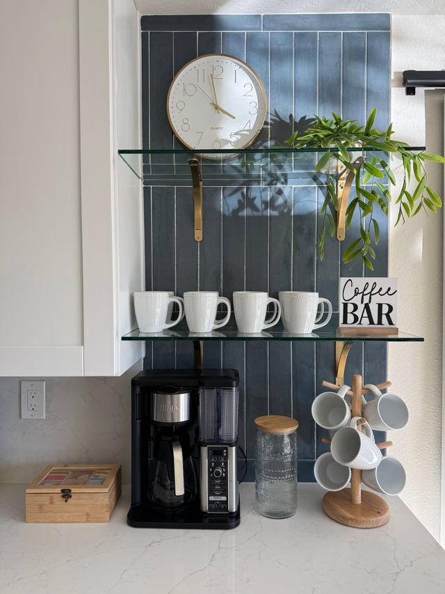 Cozy kitchen coffee station with navy vertical tile backsplash, round wall clock, glass shelves holding four white mugs, trailing green plant and a "Coffee Bar" sign above a black drip coffee maker and wooden mug tree on a white quartz countertop.