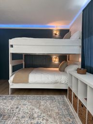 White wooden bunk bed in a cozy modern bedroom with navy wood-paneled accent wall, warm rattan wall sconces, blue LED cove lighting, woven-basket storage, neutral linens and patterned area rug.