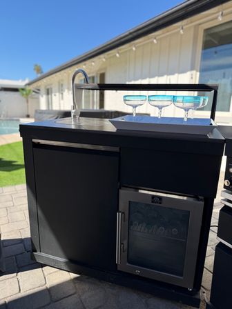 Poolside backyard patio outdoor bar with built-in sink and mini fridge, white tray holding three turquoise-rimmed coupe glasses, single-story house and blue sky in background.