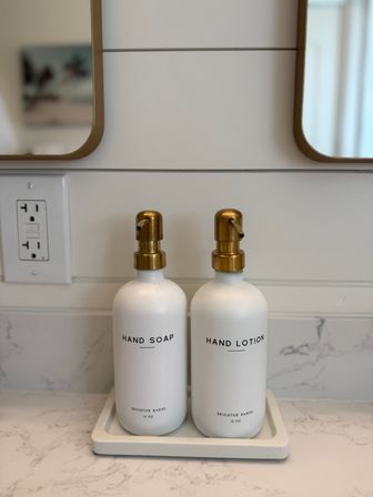 Minimal bathroom vanity scene with two white pump bottles labeled "Hand Soap" and "Hand Lotion" topped by brass dispensers on a white marble tray, shiplap wall and mirrors in the background.