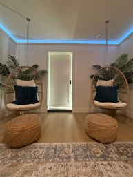 Modern boho indoor lounge with two hanging rattan egg chairs and navy pillows, woven poufs on a light wood floor, palm plants and blue LED cove lighting