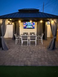 Cozy evening backyard gazebo with outdoor dining table and six chairs, wall-mounted TV, string lights and curtains over a stone paver patio