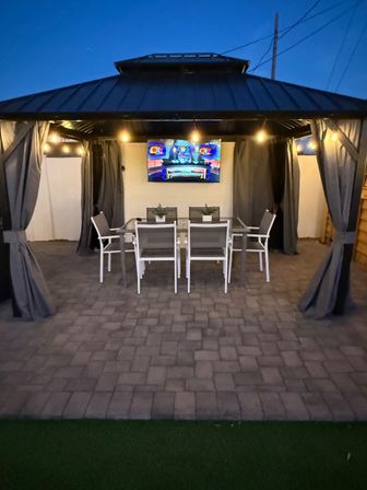 Cozy evening backyard gazebo with outdoor dining table and six chairs, wall-mounted TV, string lights and curtains over a stone paver patio