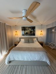 Cozy coastal bedroom with king-size bed, neutral linens, wooden ceiling fan, woven pendant lights, palm beach artwork on shiplap wall, bedside tables and striped rug — bright beachy vibe.